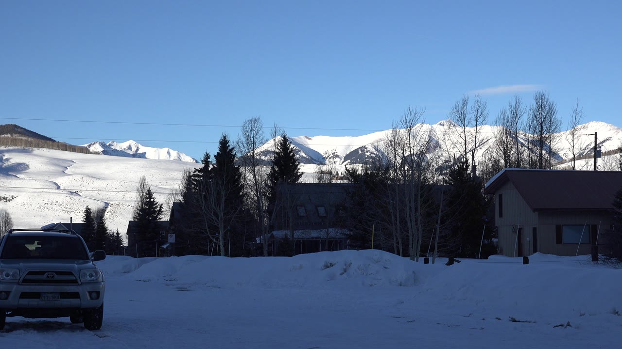 At 0:01, the pickup truck is more centered in the snowy foreground, with Crested Butte houses and trees in shadow below sunlit mountain slopes and no visible people.