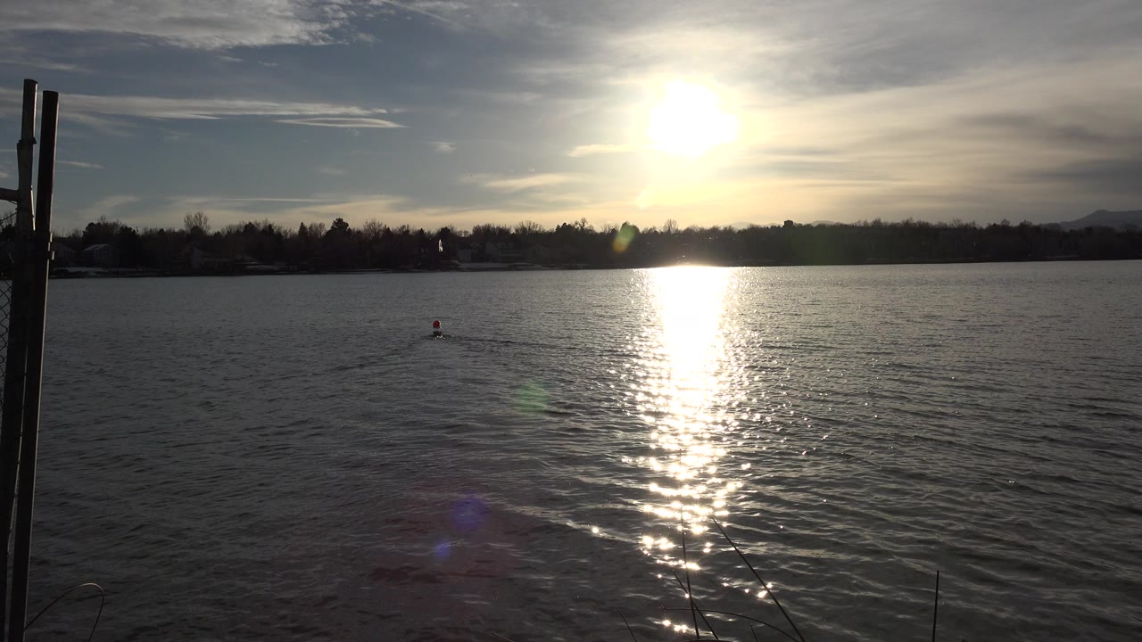 A duck swims in the midground of a Westminster, Colorado lake as the low sun reflects in a bright vertical path across rippled water, with fence posts at the left edge and no visible people.