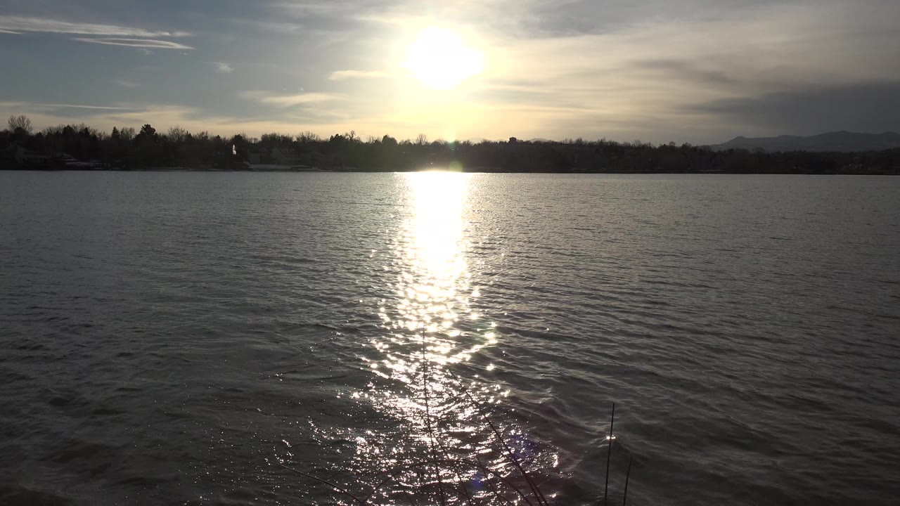 Low sun reflects across rippled lake water in Westminster, Colorado, with a dark shoreline and distant hills under a hazy sky, no visible people.