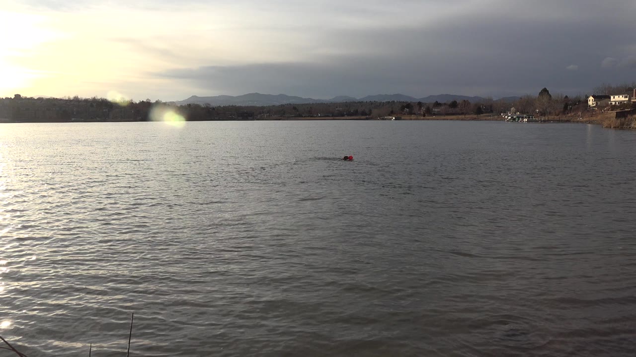 At 0:01 in Westminster, Colorado, the red and dark distant shape appears slightly farther across the lake, with textured water, a low shore, hills, bright cloud cover, and no visible people.