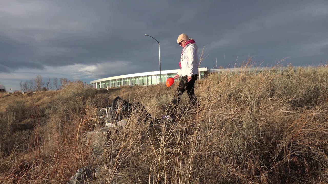Woman in white jacket stands among tall grass with dog near rocks at her feet.