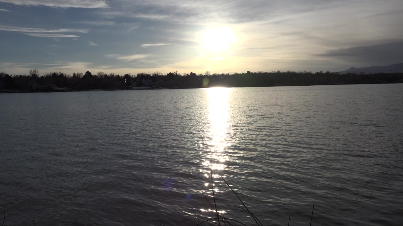 At 0:00 in Westminster, Colorado, a low sun casts a bright vertical reflection across rippled lake water, with a dark tree-lined shore and no visible people.