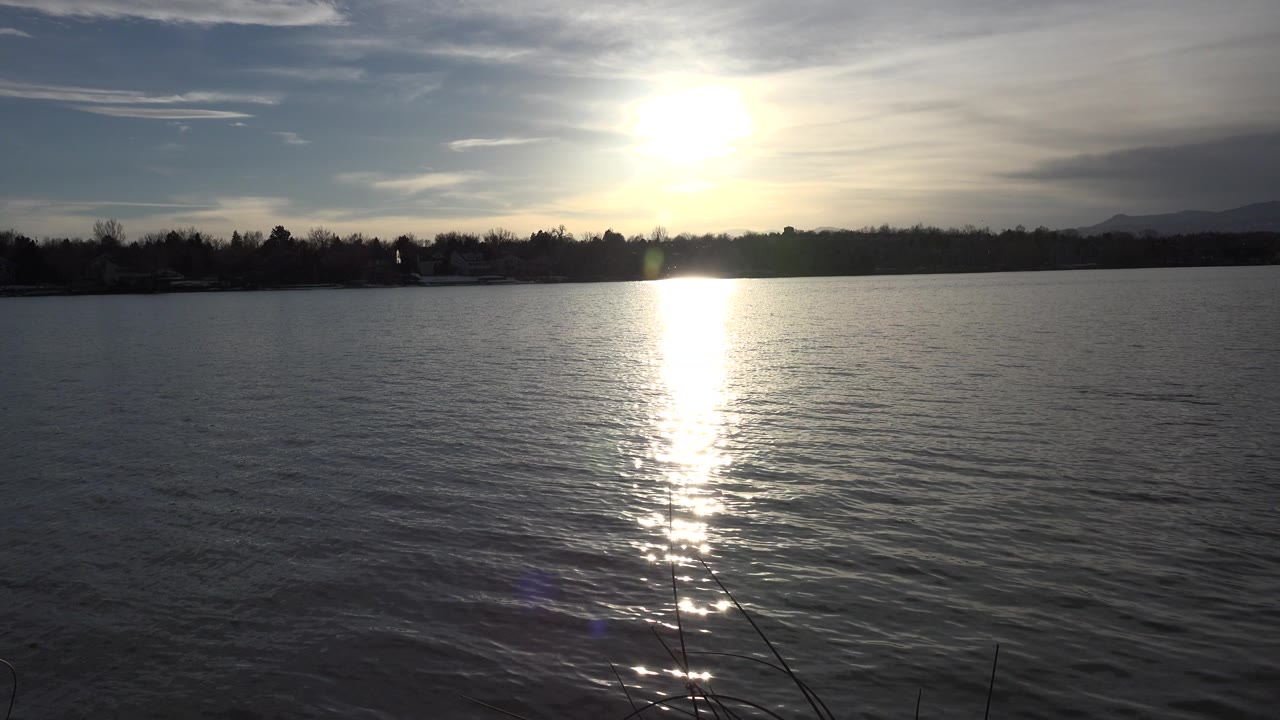At 0:01 in Westminster, Colorado, the sun glare remains centered over the lake while small ripples break the reflection, with silhouetted trees and no visible people.