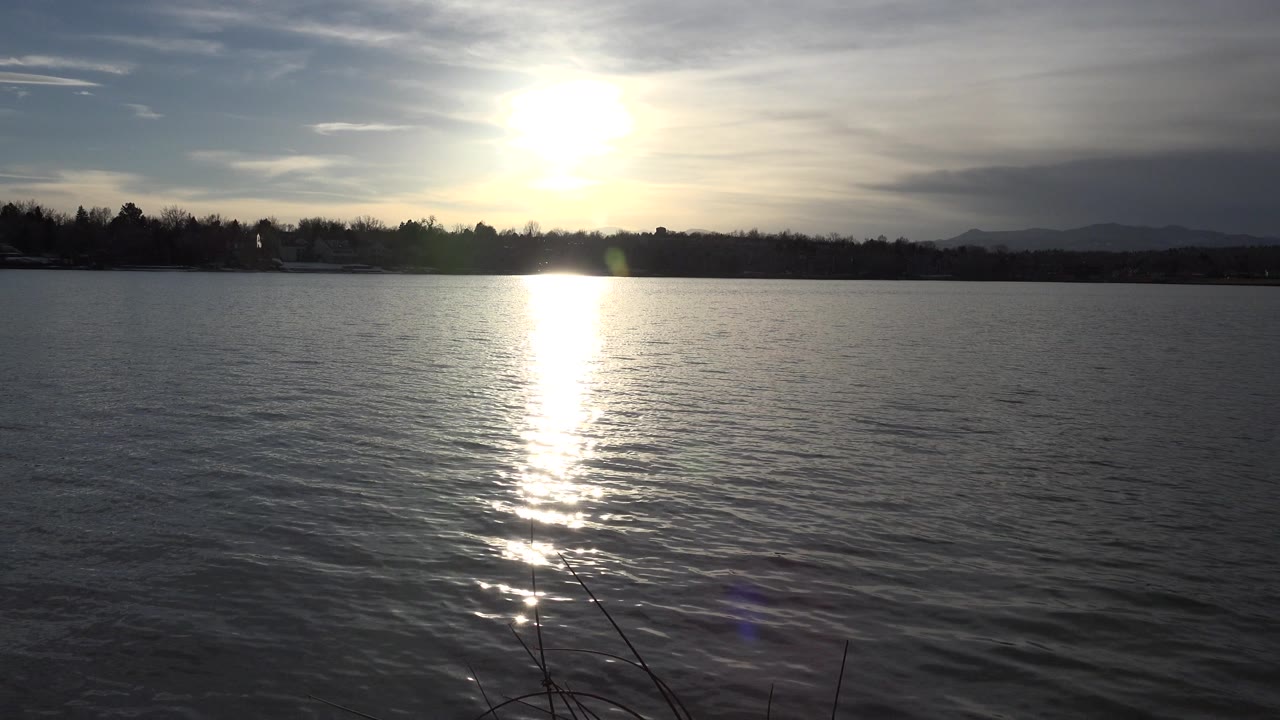 Low sun reflects in a bright vertical streak across a Westminster, Colorado lake, with rippled water, dark shoreline, distant hills, foreground reeds, and no visible people.