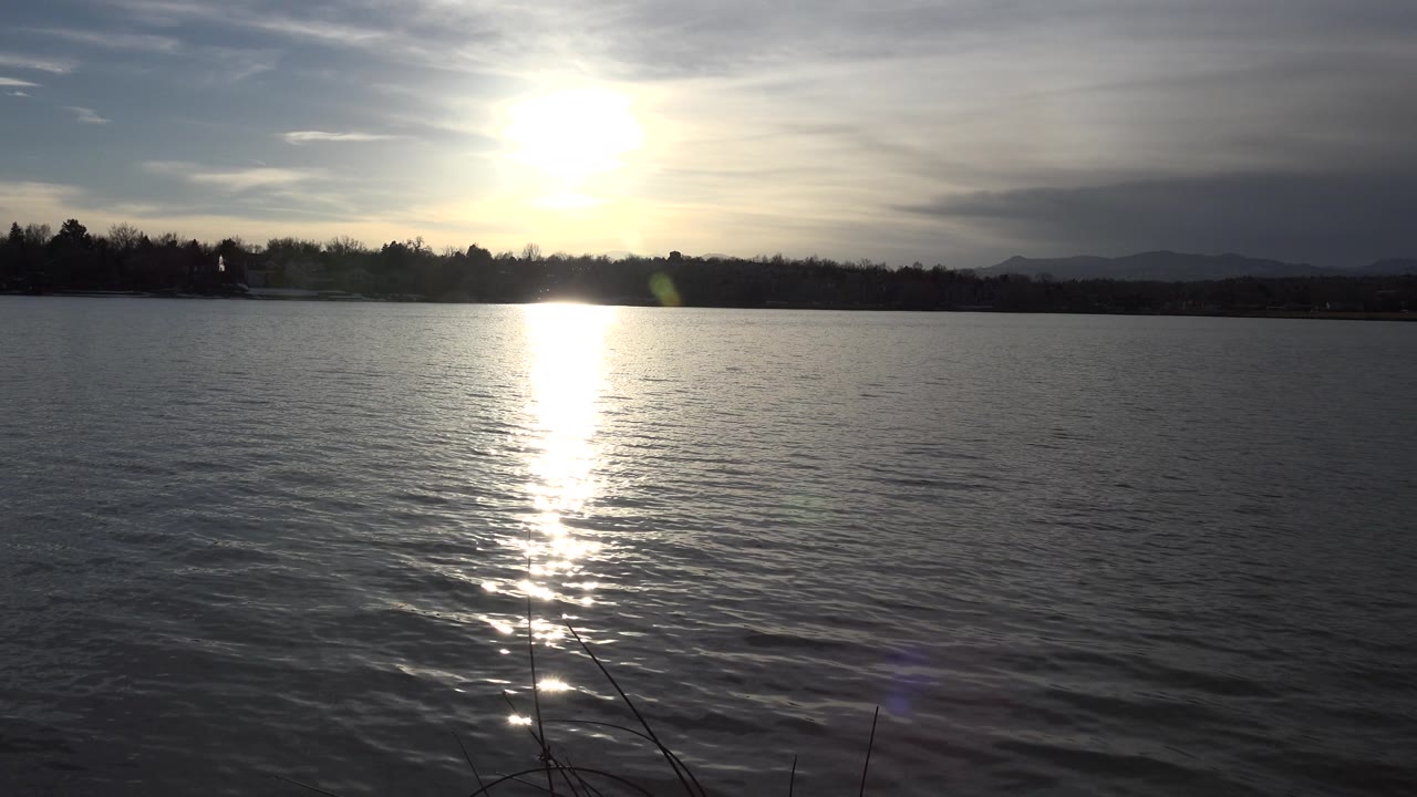 Sunlit lake with a dark tree line, distant hills, and small lens flare near the water.