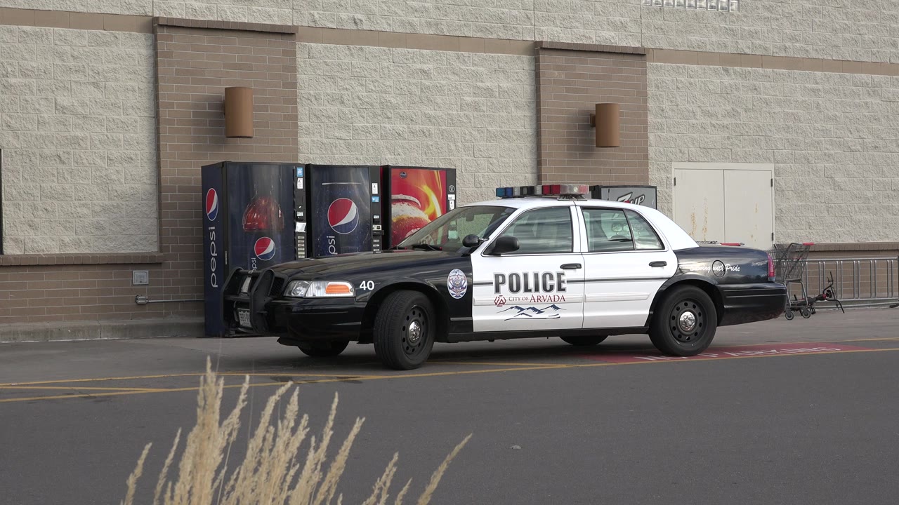 A black-and-white police car is parked at the curb outside a Westminster, Colorado store, with Pepsi vending machines behind it, dry grasses in the foreground, and no visible people.