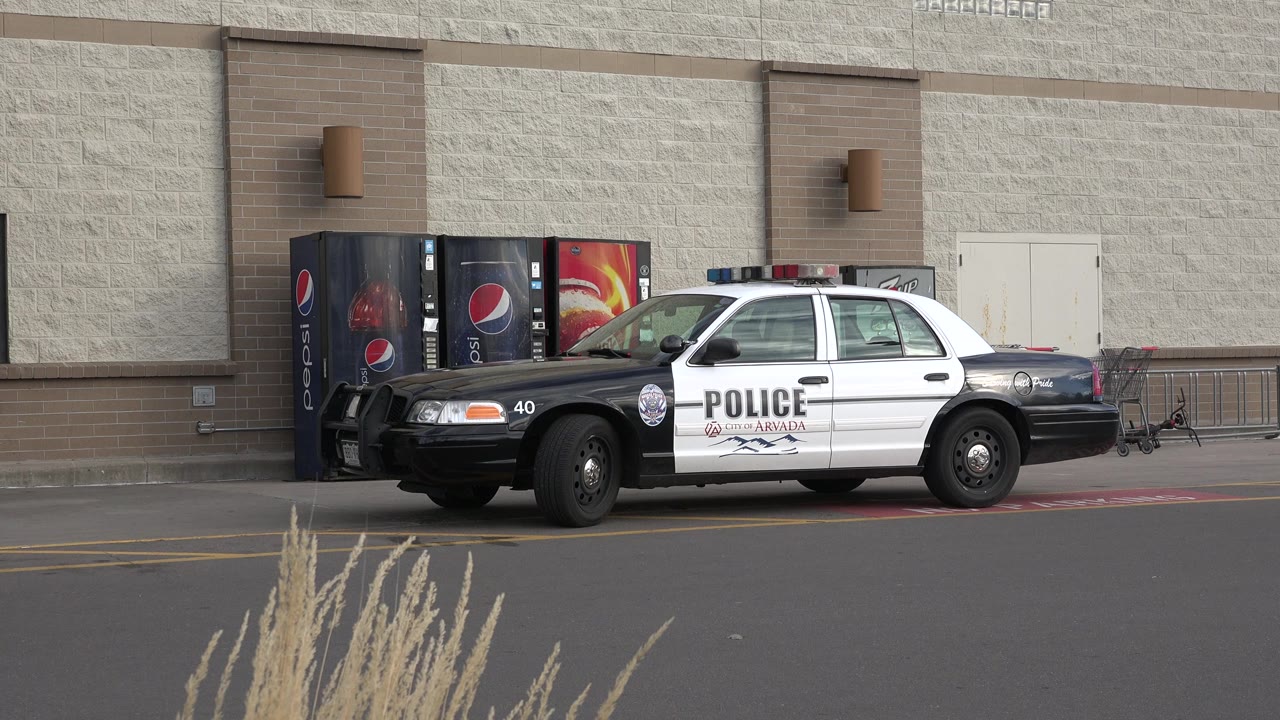 The parked police sedan remains centered outside the Westminster, Colorado retail wall, with vending machines, a small window, curb markings, and no visible people.