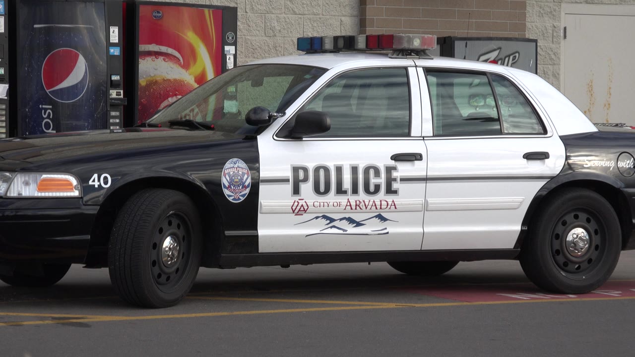 The police car remains parked in profile outside the building in Westminster, Colorado, with Pepsi vending machines and a plain wall behind it and no visible people.