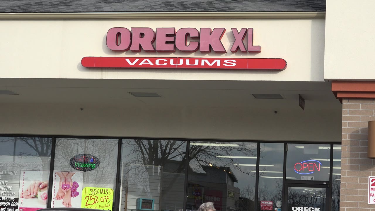 Oreck XL Vacuums storefront with window posters and parked cars.