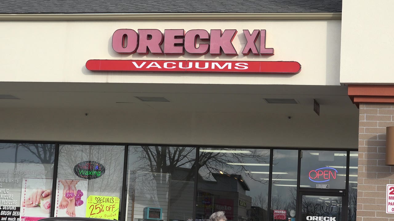 The Westminster storefront remains framed straight on, with the Oreck XL Vacuums sign near the top, reflected trees in the windows, bright sale posters, and no clearly visible people.