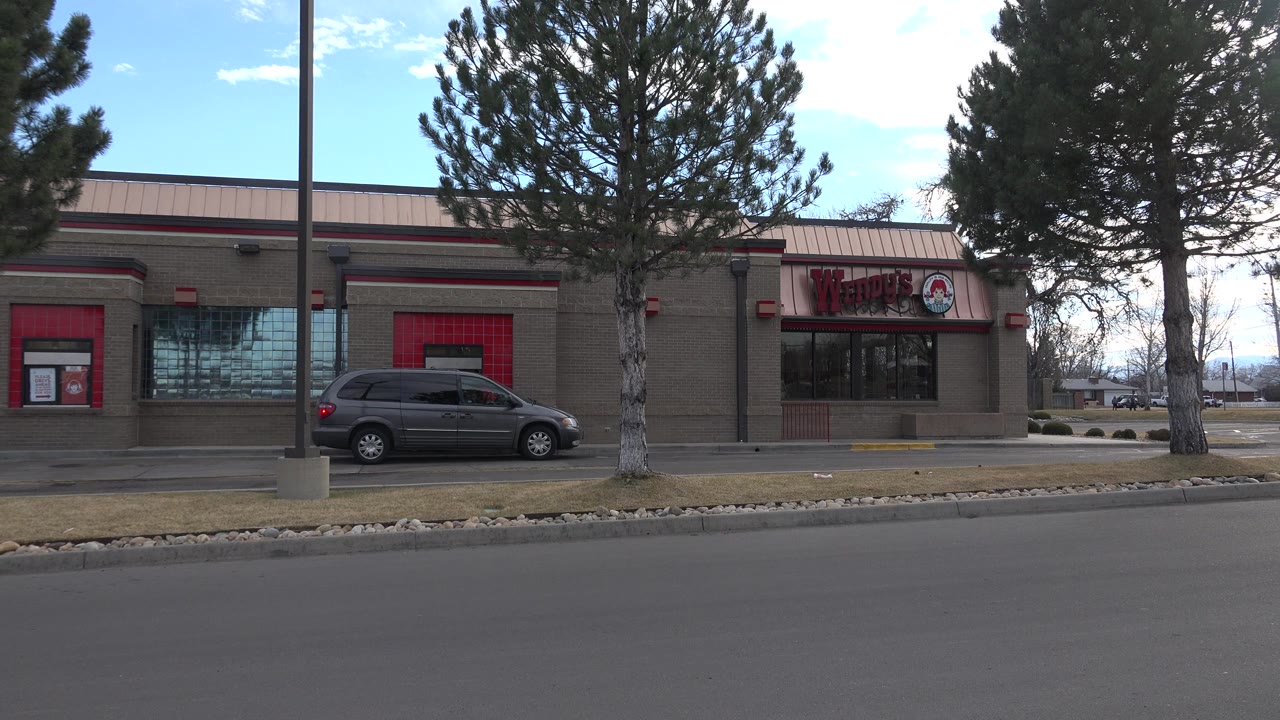 A gray van waits near the Wendy's drive-thru window in Westminster, Colorado, with pine trees, a lamppost, and an empty asphalt lane in front, with no clearly visible people.