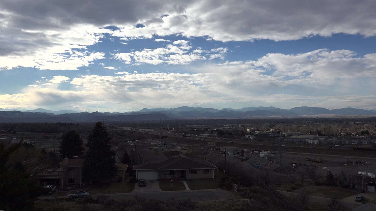 Westminster, Colorado suburban foreground with rooftops, trees, a lightly traveled road, distant mountain silhouettes, bright cloud breaks, and no visible people.