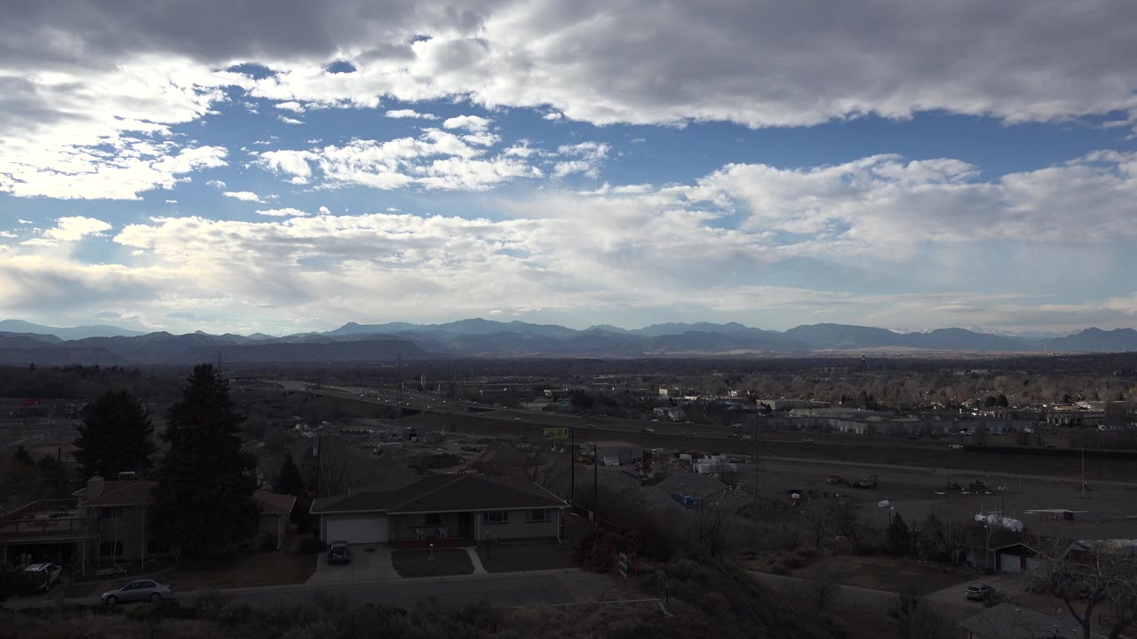 Westminster, Colorado view with a shadowed residential foreground, open midground roads and lots, blue sky openings, layered clouds, mountain horizon, and no visible people.