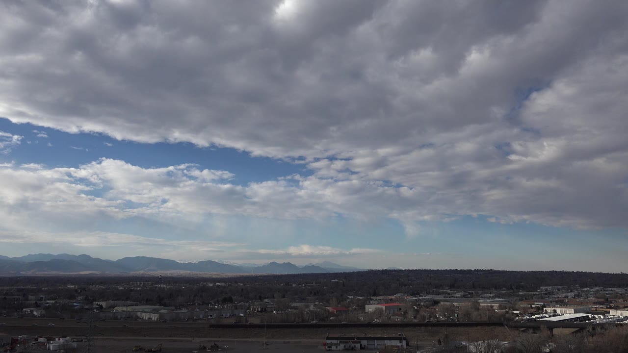 Westminster, Colorado frame with a broad blue opening under billowing clouds, a dark town strip, and distant mountain silhouettes, with no visible people.