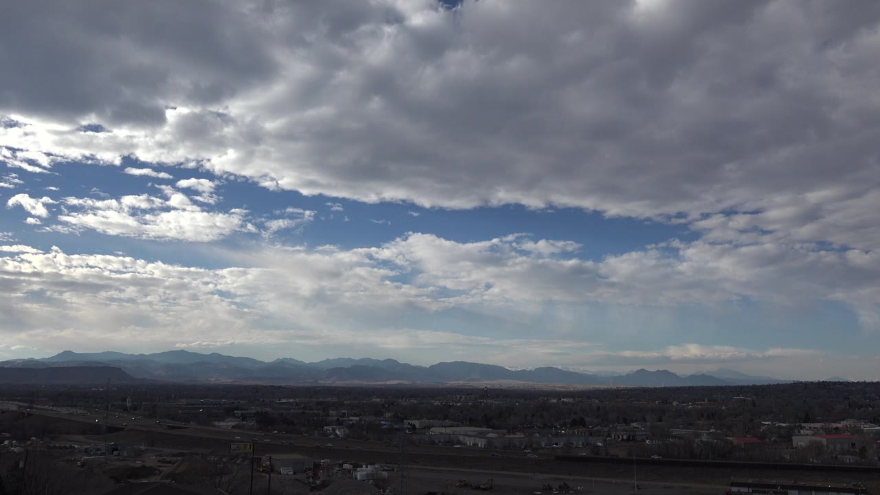 Westminster, Colorado panorama with layered gray clouds, sunlit cloud breaks, a low dark town foreground, distant mountains, and no visible people.