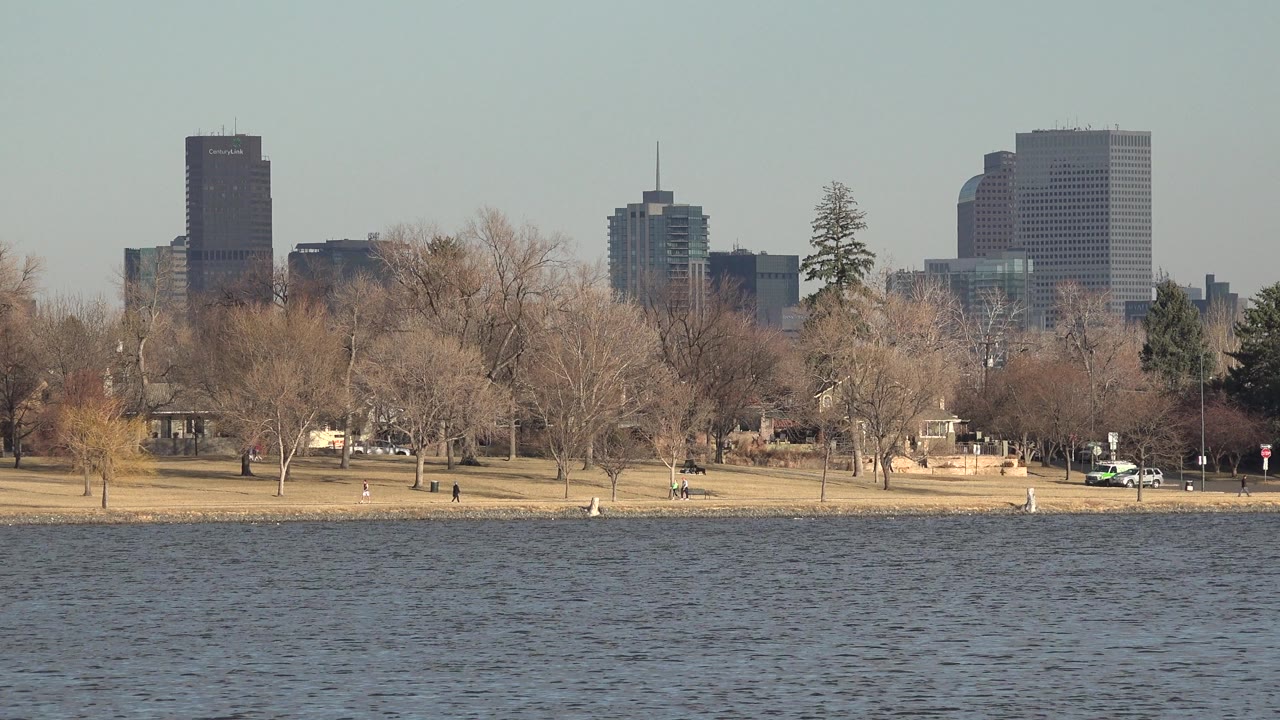Westminster, Colorado lakefront view with textured water in front, scattered park trees, tiny walkers along the shore, and high-rise buildings in the distance.