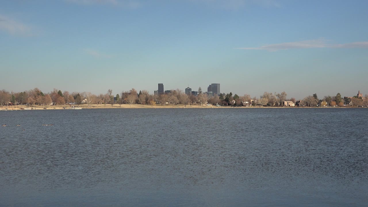 Wide Westminster, Colorado water view with rippled lake foreground, a thin shoreline of trees, distant buildings on the horizon, blue sky above, and no visible people.