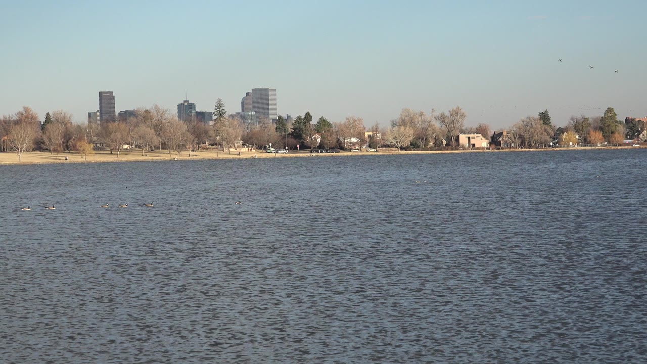 Opening frame of a Westminster, Colorado lake view with rippled water in front, bare shoreline trees, distant high-rise buildings, and no visible people.