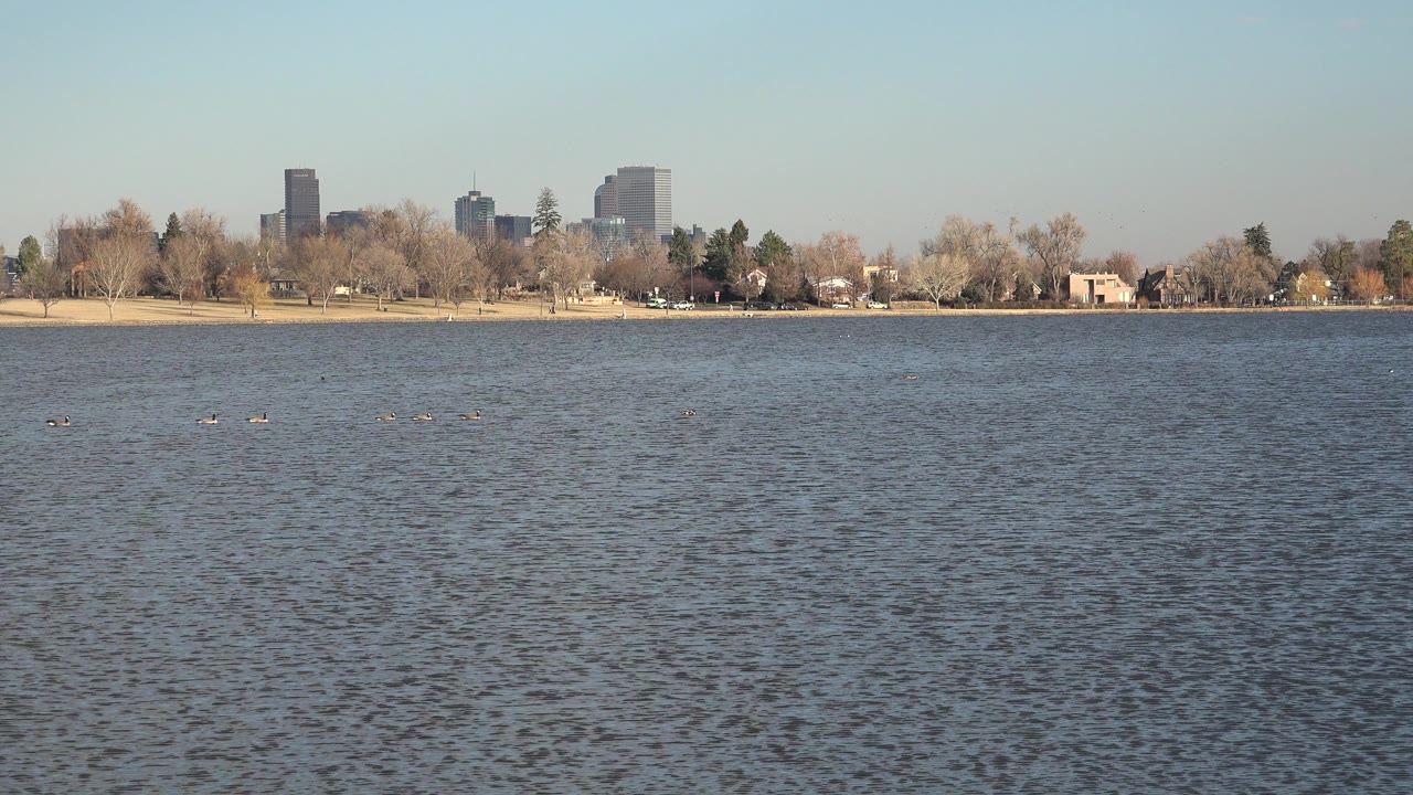 Westminster, Colorado shoreline seen across textured water, with leafless trees, low park structures, taller buildings in back, and no visible people.