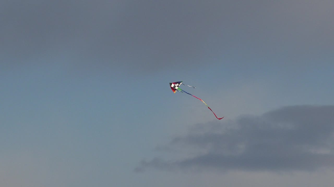 At 0:00 in Westminster, Colorado, a small colorful kite with a red trailing tail sits high against blue sky above a soft gray cloud band, with no visible people.