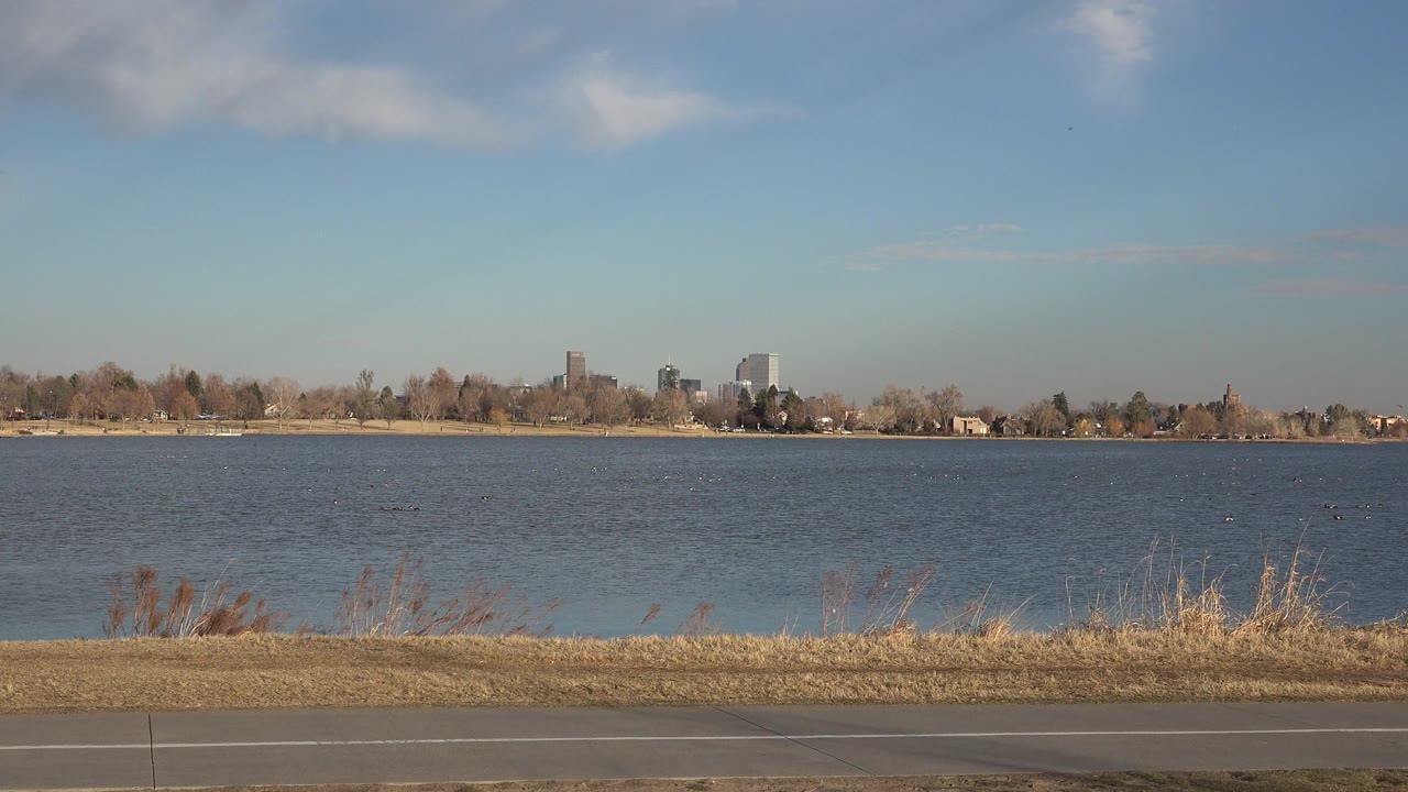 Wide water view with dry grasses, path, trees, and distant buildings.