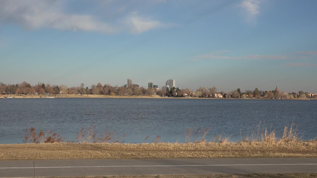 Westminster, Colorado shoreline frame showing rippled water, tawny grasses, a paved path along the bottom edge, distant trees and buildings, and no visible people.