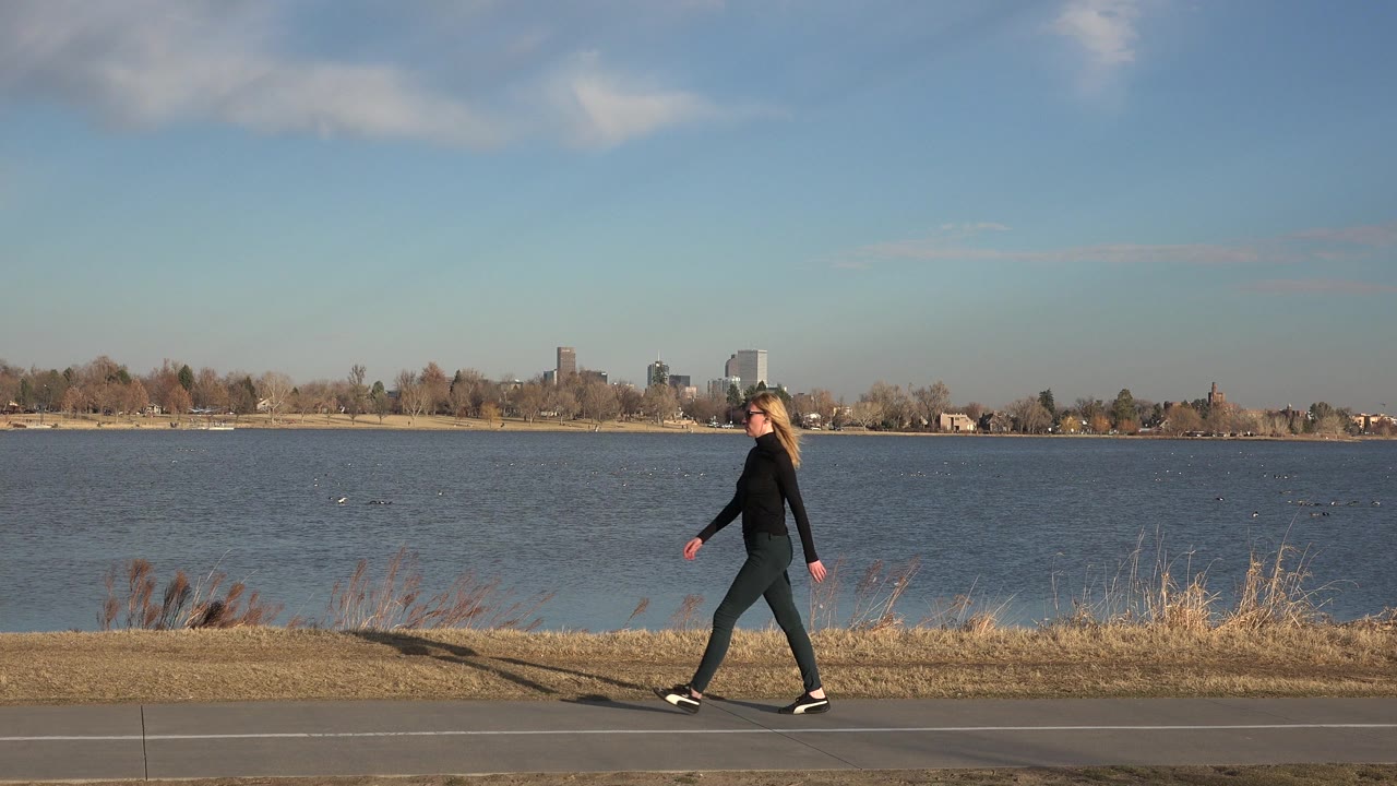 The woman continues walking beside the water in Westminster, Colorado, with her shadow on the pavement and a low skyline across the far shore.
