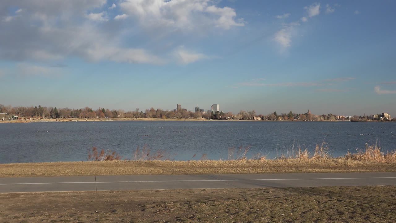 Wide lakeside view with path, dry grass, water, distant trees, and clouds.
