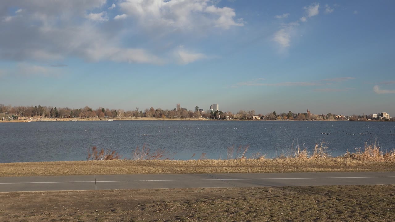 The Westminster lakeside remains empty, with the path and brown grass across the foreground, a broad band of lake water, and a low tree line beneath scattered clouds.