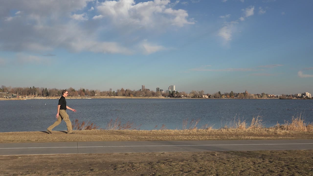 A man in a black shirt walks along a paved road beside a lake in Westminster, Colorado, with dry grass and a distant shoreline behind him.