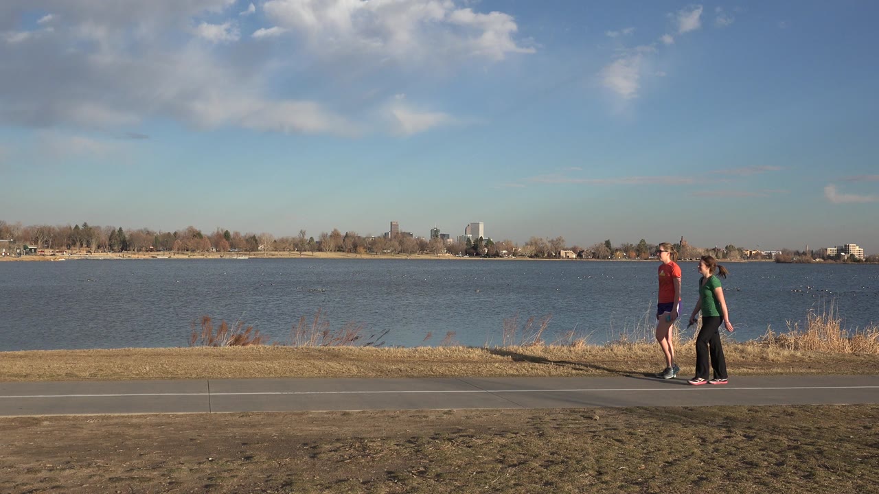 Two people walking on a paved lakeside path with water and distant buildings behind them.