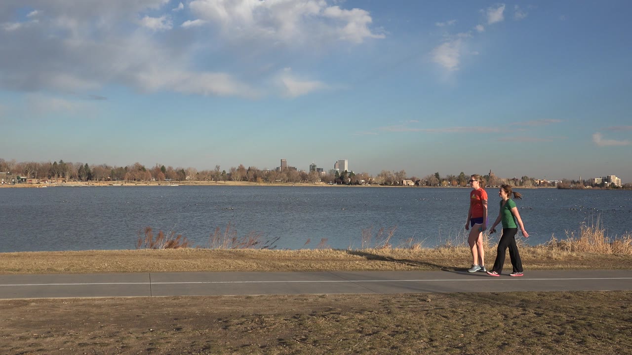 Two people continue across the lower frame on a lakeside path in Westminster, Colorado, with blue water filling the midground and scattered clouds above.