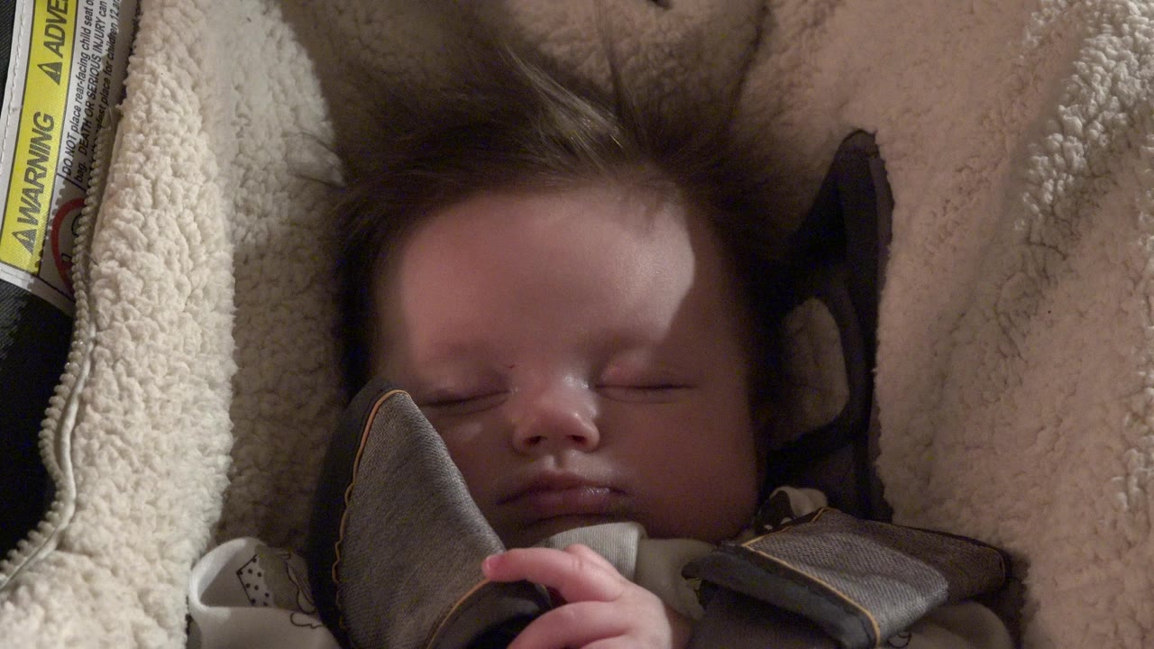 At 0:00, an infant sleeps with eyes closed in a padded car seat in Westminster, Colorado, with cream fleece lining and a yellow warning label visible at the left edge.