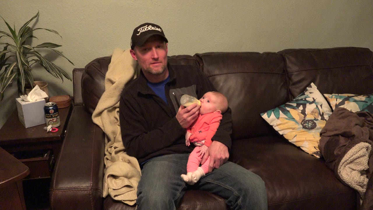 At the start of the Westminster, Colorado clip, a father sits centered on a brown leather couch and holds a baby while feeding from a bottle in a warmly lit living room.