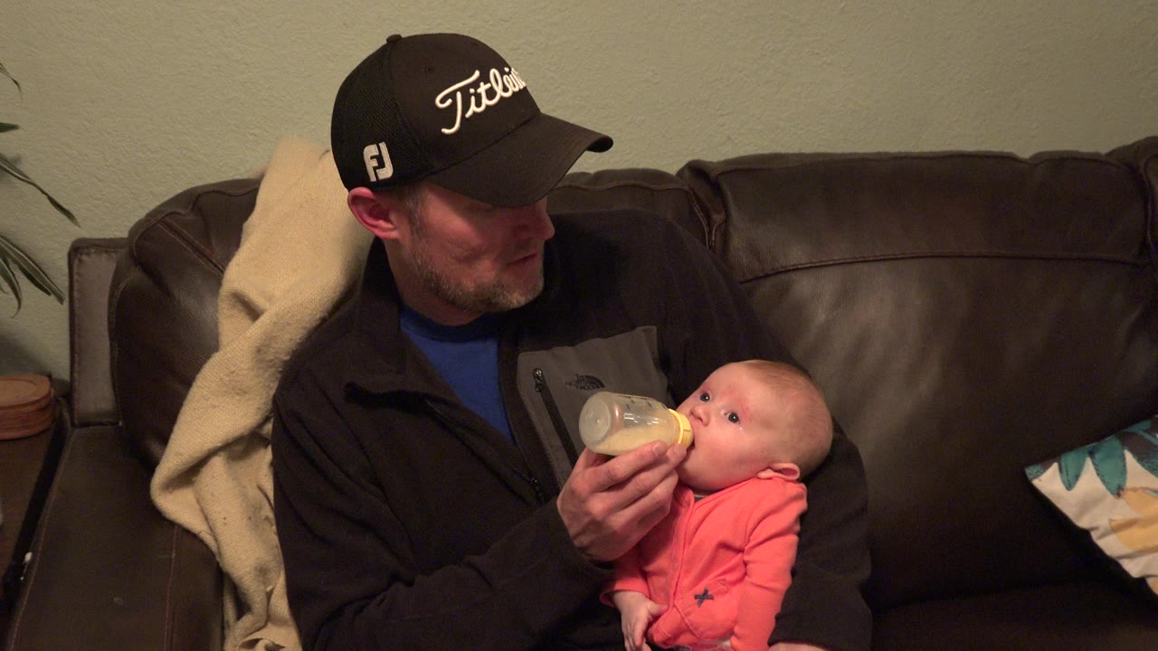 In Westminster, Colorado, a father in a black cap looks down while holding a bottle for a baby seated on his lap on a dark leather sofa.