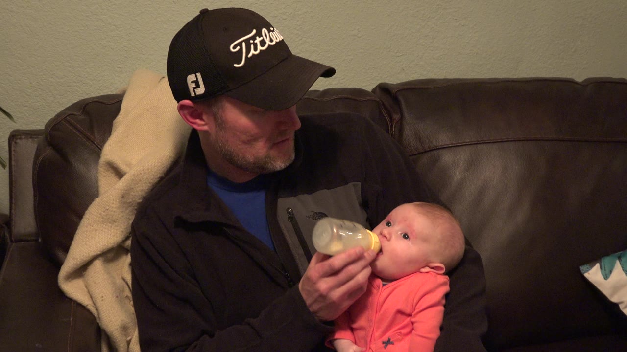 Man in cap feeding baby with bottle on couch.