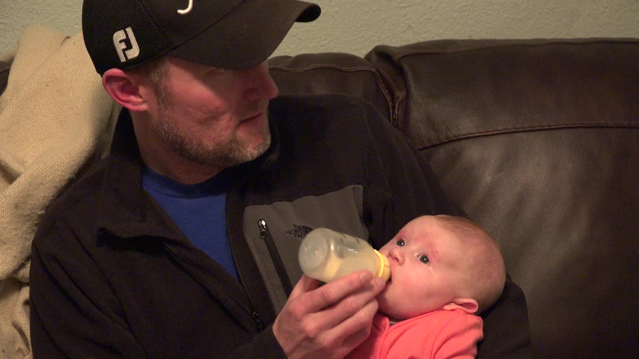 Father in a black cap bottle-feeding a baby on a leather couch.