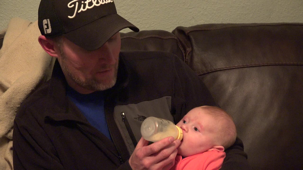 In Westminster, Colorado, a man in a dark cap looks down while feeding a baby from a bottle on a brown leather sofa.
