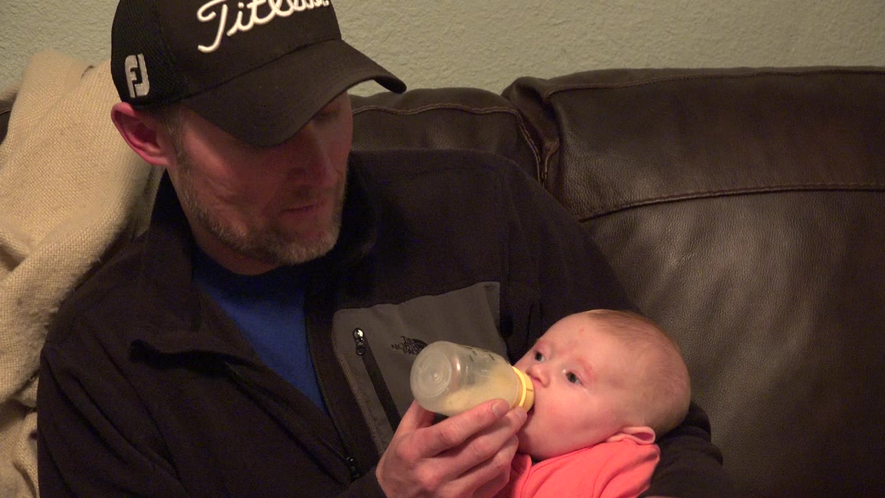 The man keeps the bottle at the baby's mouth while sitting close on a brown sofa in the Westminster, Colorado living room.