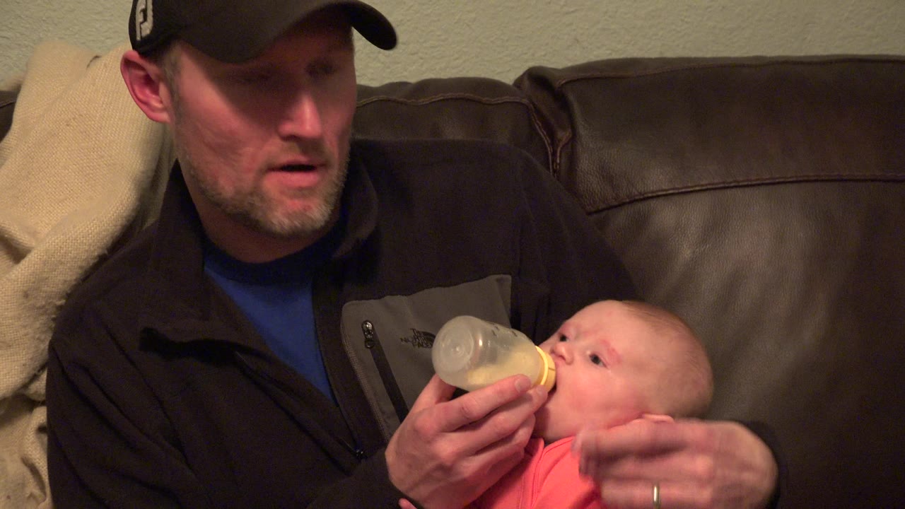 Man adjusts a baby while holding a bottle on a couch.