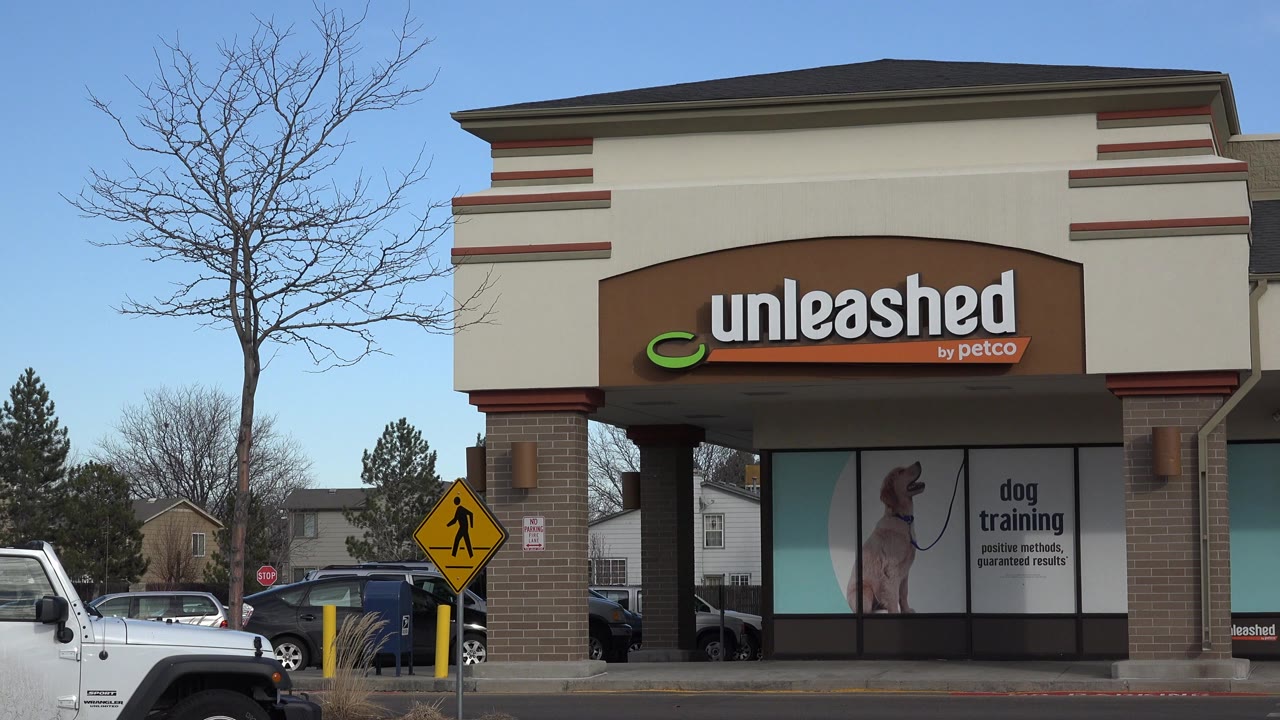 Opening frame of an Unleashed by Petco storefront in Westminster, Colorado, with a bare tree, pedestrian crossing sign, parked cars, and no visible people.