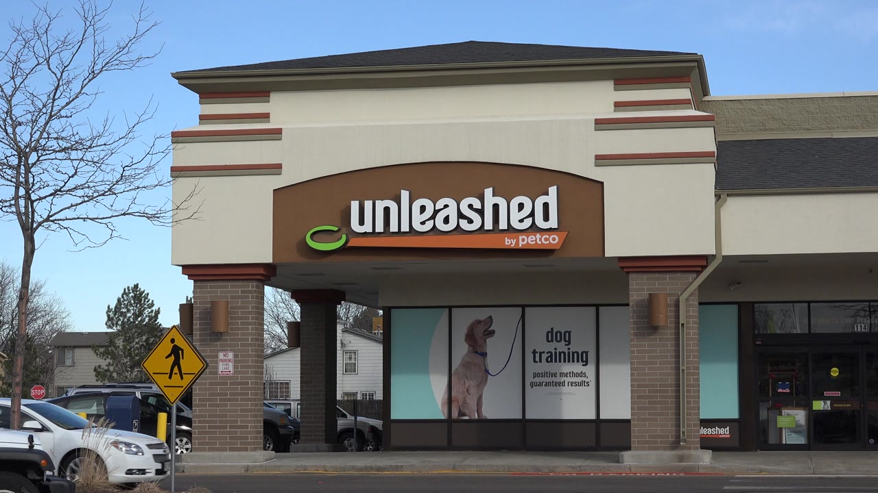 Westminster, Colorado strip mall storefront remains centered as a white car begins crossing the parking lot edge at frame right, with no visible people.