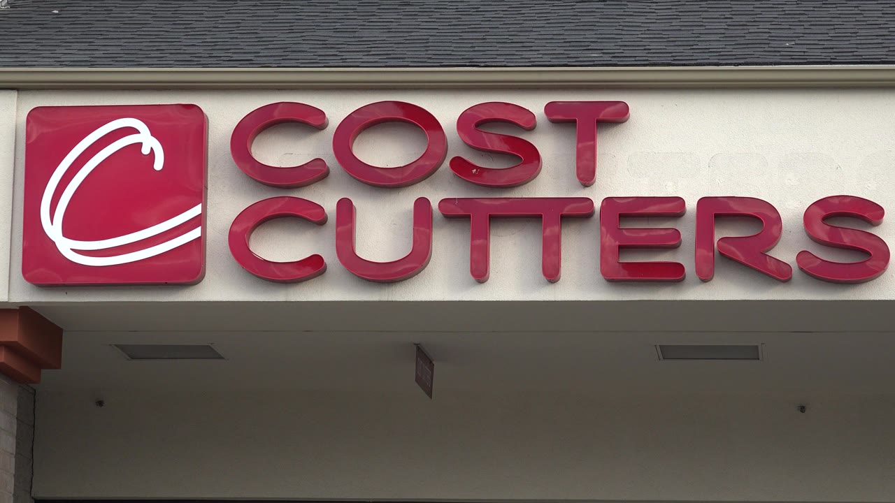 Tight opening frame of the red Cost Cutters logo and lettering on a white wall in Westminster, Colorado, with roof shingles above and no visible people.