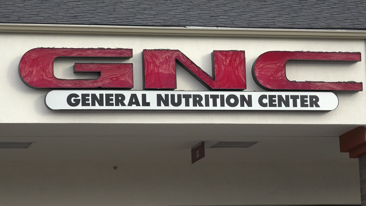 Opening frame of a red GNC General Nutrition Center sign on a beige storefront in Westminster, Colorado, with roof shingles above and no visible people.
