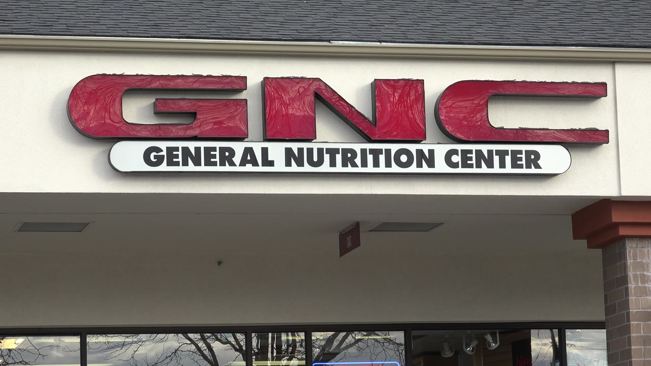 Close view of a red GNC General Nutrition Center sign on a beige storefront.