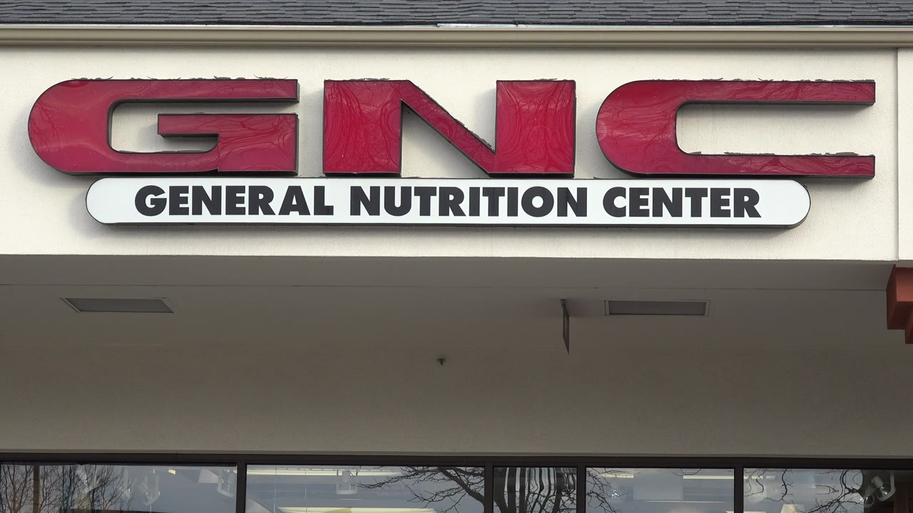The red GNC sign remains centered over a beige storefront overhang in Westminster, Colorado, with window glass and bare tree reflections below and no visible people.