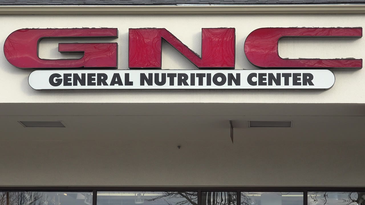 Opening frame of a red GNC General Nutrition Center sign on a beige storefront in Westminster, Colorado, with window reflections below and no visible people.