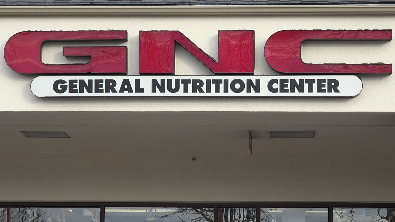 A tight Westminster storefront view centers the red GNC letters and white General Nutrition Center panel above dark-framed windows, with no visible people.