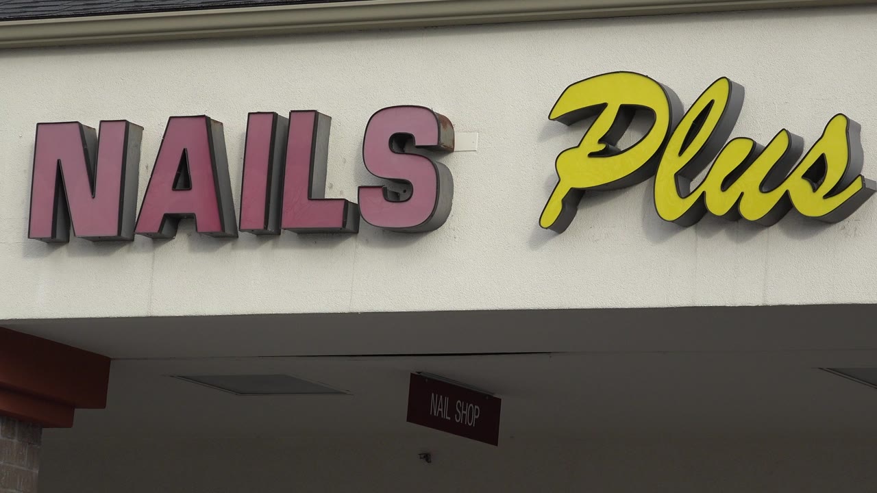 Close opening frame of the Nails Plus storefront sign in Westminster, Colorado, with red block letters, yellow script, beige facade, and no visible people.