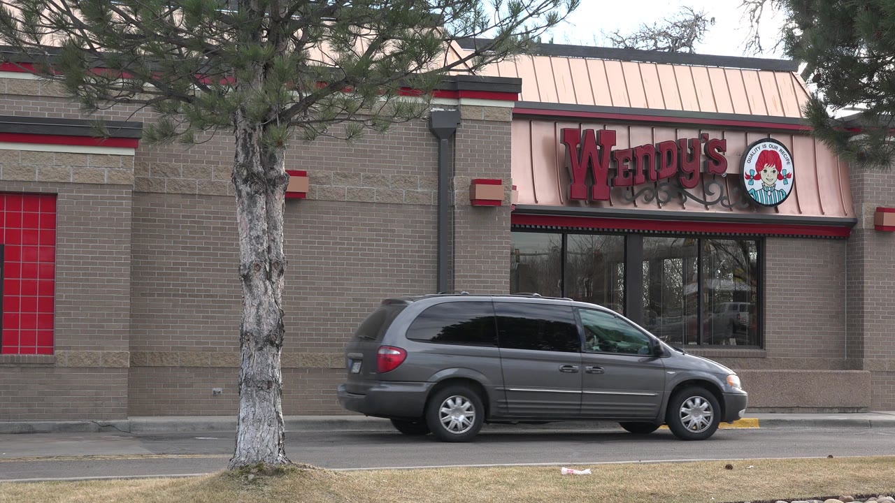 Wendy's facade in Westminster, Colorado, with red signage above dark windows, a gray minivan along the curb, foreground trees, and no visible people.
